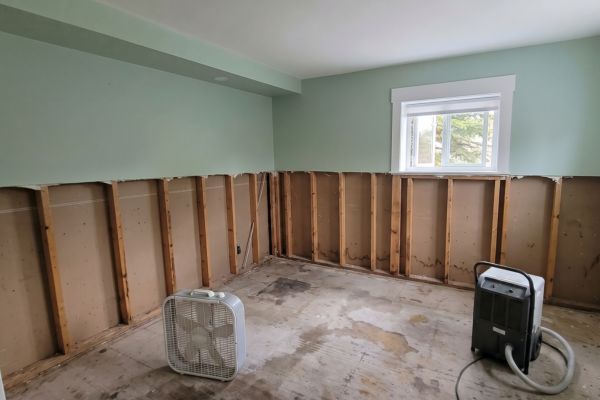 Bare wall studs in a flood-damaged room with a box fan and dehumidifier drying the space